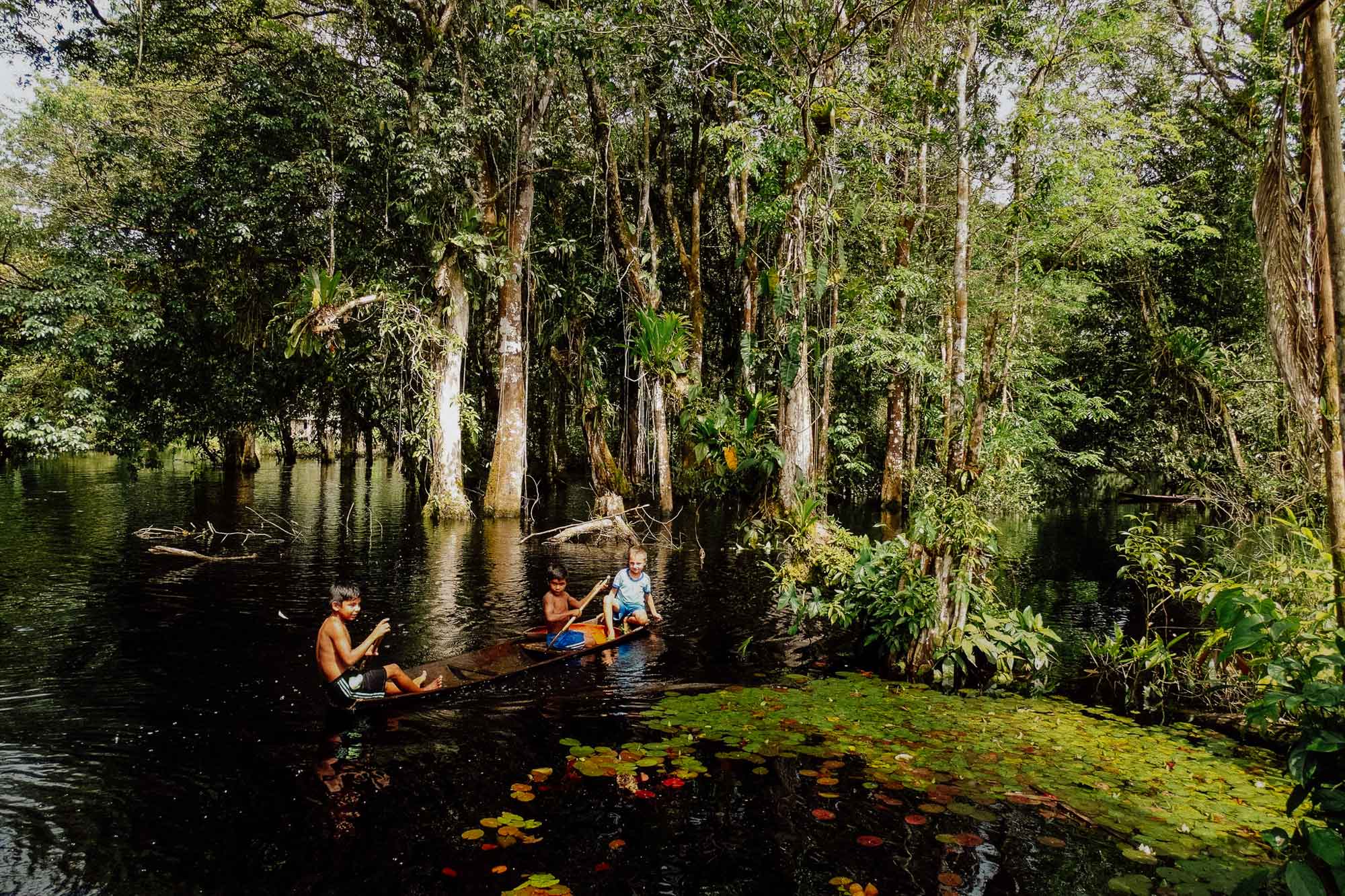 Drei Kinder in einem Kanu auf einem Fluss. Im Hintergrund ist Dschungel zu sehen.
