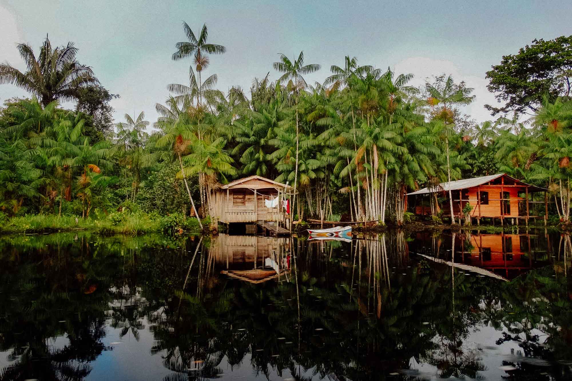 Zwei Hütten am Flussufer mit Palmen im Hintergrund.